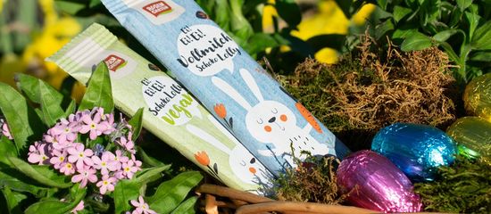 Eine Nahaufnahme auf einen Osterkorb neben lila Blumen. Mit Schoko-Lollys und Schokoeiern in bunter Folie.
