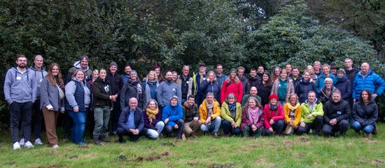 Eine große Gruppe von Menschen steht zusammen auf einer Wiese vor Bäumen.