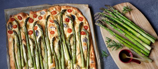 Focaccia mit Spargel und Tomaten auf Holzplatte, daneben frischer grüner Spargel und Rosmarin.