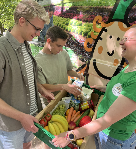 KI generiert: Drei Personen begutachten eine Kiste mit frischem Obst und Gemüse vor einem bunt bemalten Lieferwagen.