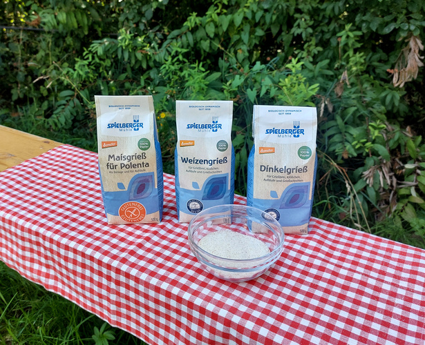 KI generiert: Drei Packungen Grieß (Mais-, Weizen-, Dinkelgrieß) auf karierter Tischdecke, mit einer Schale Grieß.