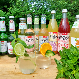 KI generiert: Verschiedene BioZisch-Getränkeflaschen und Gläser mit Limette und Orange auf einem Holztisch im Freien.