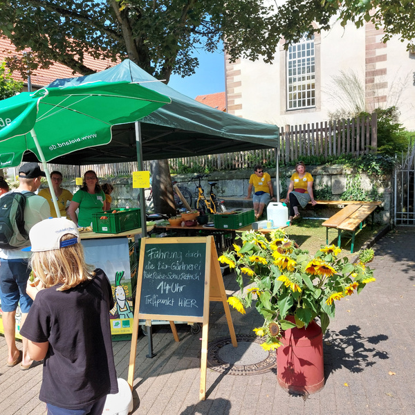 KI generiert: Ein Marktstand mit Leuten, Sonnenblumen und einem Schild. Text: "Führung durch die Bio-Gärtnerei... 14 Uhr".