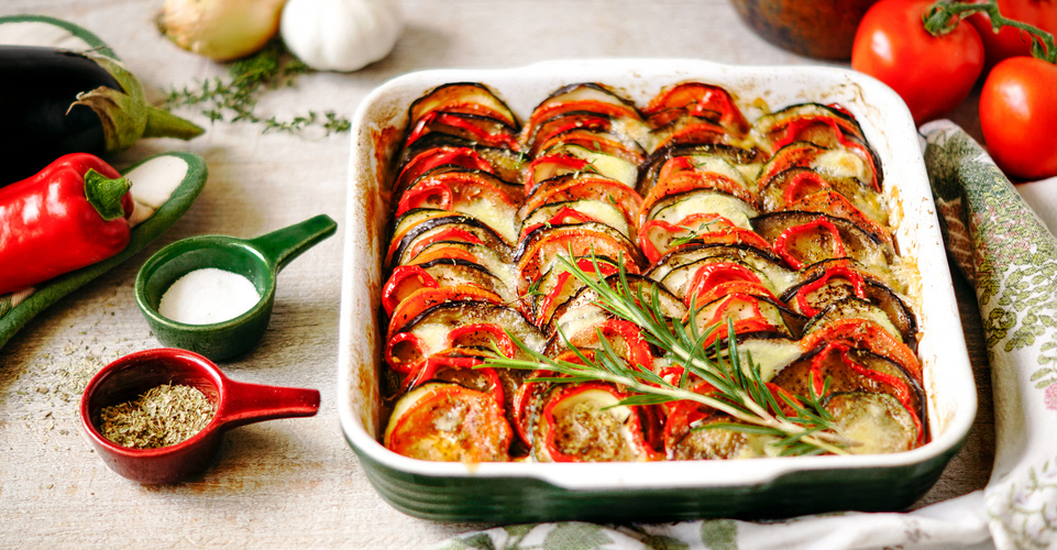 KI generiert: Ein Gemüseauflauf mit Tomaten, Zucchini und Auberginen in einer Backform, umgeben von Kräutern und Gewürzen.