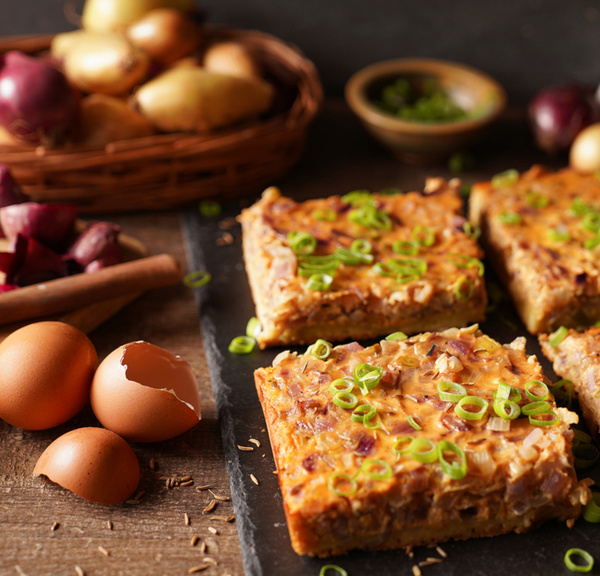 KI generiert: Quadratische Zwiebelkuchenstücke mit grünen Zwiebeln, daneben Eier und Zwiebeln auf einem Tisch.