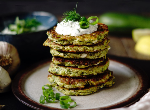 KI generiert: Ein Stapel Zucchini-Puffer mit Kräuterdip auf einem Teller, dekoriert mit Frühlingszwiebeln und Dill.