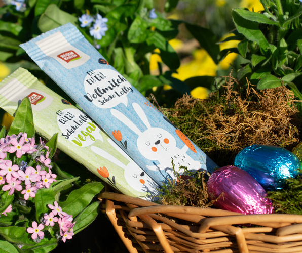 KI generiert: Ein Osterkorb mit Schoko-Lollys und Schoko-Eiern, umgeben von Blumen. Text: "Vollmilch Schoko Lolly vegan".