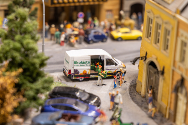 KI generiert: Ein Lieferwagen der "Ökokiste" in einer Miniaturstadt; Menschen interagieren. Text: "www.oekokiste.de".