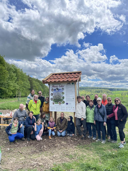 KI generiert: Eine Gruppe von Menschen versammelt sich im Freien um ein großes Schild mit Informationen, wahrscheinlich zu einem Natur- oder Landschaftsprojekt. Der Hintergrund zeigt einen bewölkten Himmel und eine grüne Landschaft.