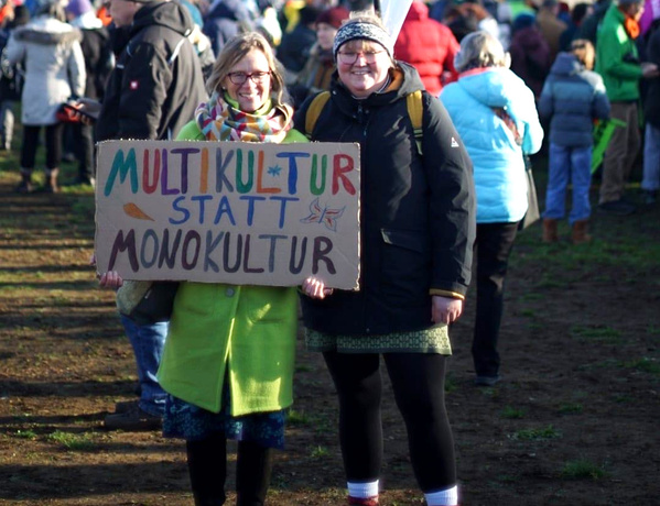  Zwei Personen halten ein Schild mit der Aufschrift: "MULTIKULTUR STATT MONOKULTUR" bei einer Versammlung.