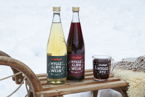 KI generiert: Zwei Flaschen Voelkel Hygge Glühwein und eine Tasse auf einem Schlitten im Schnee. Text: "Voelkel HYGG E".