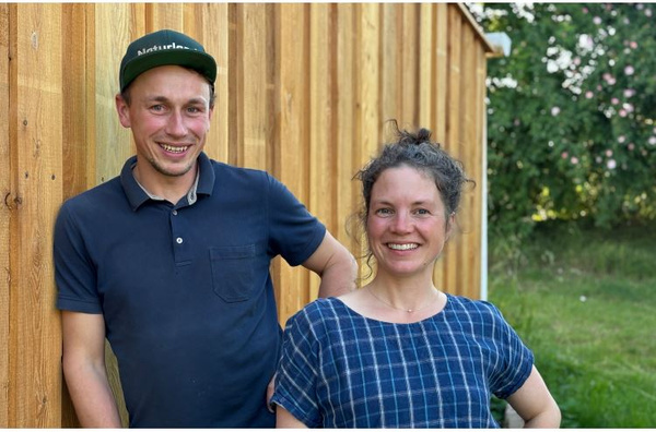 KI generiert: Zwei Menschen posieren vor einer Holzwand im Freien. Der Mann trägt ein Poloshirt und eine Mütze, die Frau ein kariertes Oberteil.