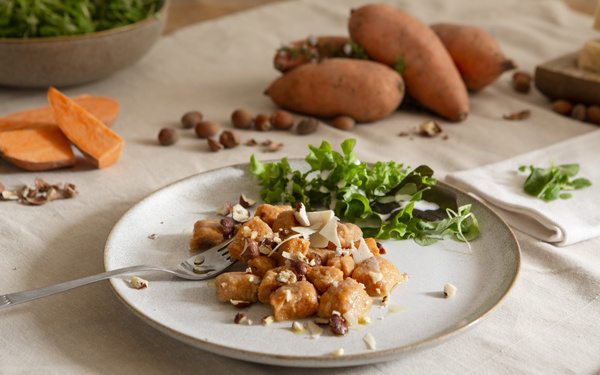 KI generiert: Ein Teller mit Gnocchi, Nüssen und Salat; im Hintergrund Süßkartoffeln.