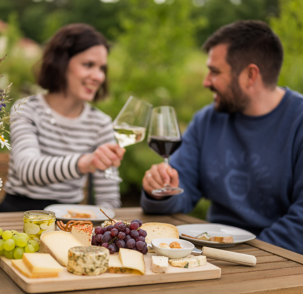 KI generiert: Ein Paar stößt zum Picknick mit Wein an, Käseplatte im Vordergrund.