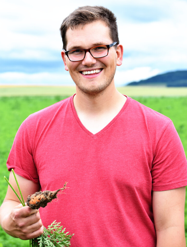 KI generiert: Ein Mann im roten Shirt hält eine frisch geerntete, erdige Karotte auf einem Feld.