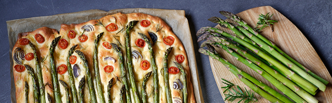 KI generiert: Spargel-Focaccia mit Tomaten und Zwiebeln auf Backpapier, daneben frischer grüner Spargel auf einem Holzbrett.