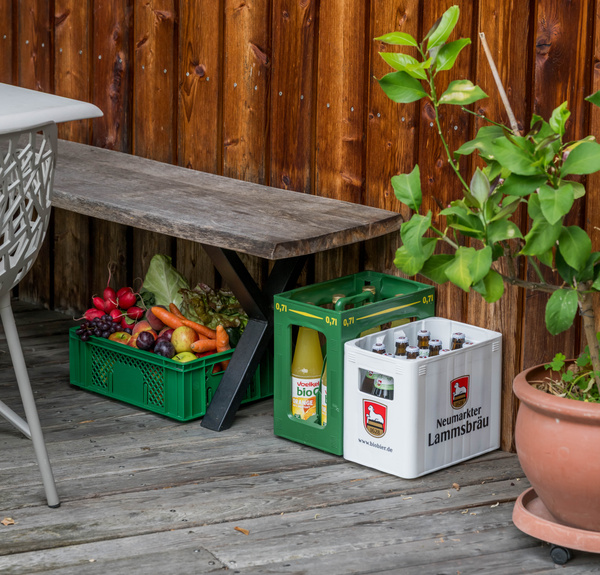 KI generiert: Gemüsekiste und Getränkekästen stehen auf einer Holzterrasse neben einer Bank und einem Blumentopf.