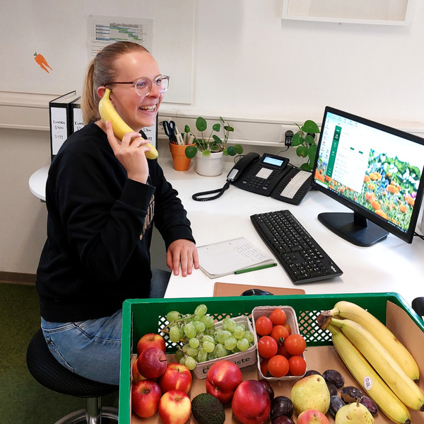 KI generiert: Eine Person telefoniert mit einer Banane. Im Vordergrund ist eine Kiste mit Obst zu sehen.