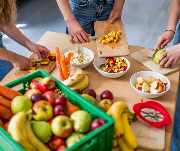 KI generiert: Personen schneiden frisches Obst auf einem Tisch; eine Kiste voller bunter Früchte steht bereit.