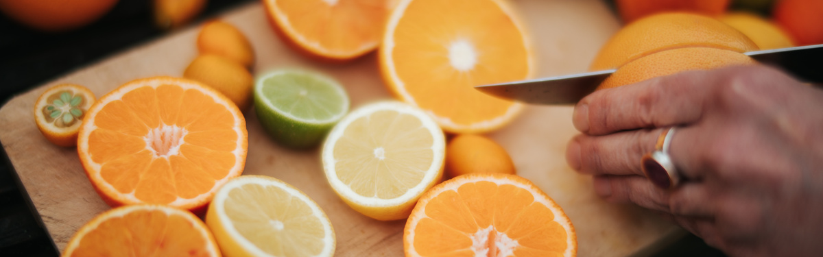 KI generiert: Auf dem Bild sind verschiedene Zitrusfrüchte auf einem Holzschneidebrett zu sehen, darunter Orangen, Zitronen und Limetten, die teilweise aufgeschnitten sind. Eine Hand schneidet mit einem Messer eine Orange.