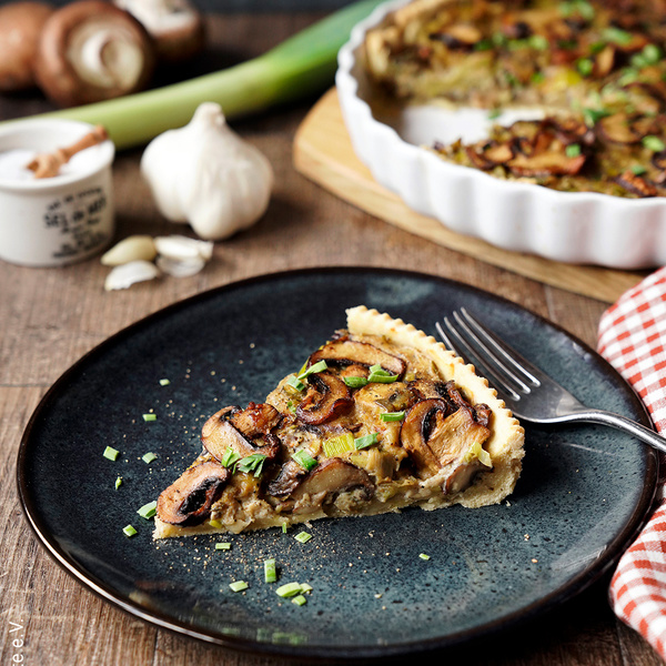 KI generiert: Ein Pilzquiche-Stück auf einem Teller, mit Besteck und Zutaten im Hintergrund.