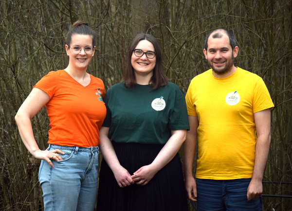 KI generiert: Drei Personen lächeln in die Kamera, sie tragen bunte T-Shirts mit einem Logo.