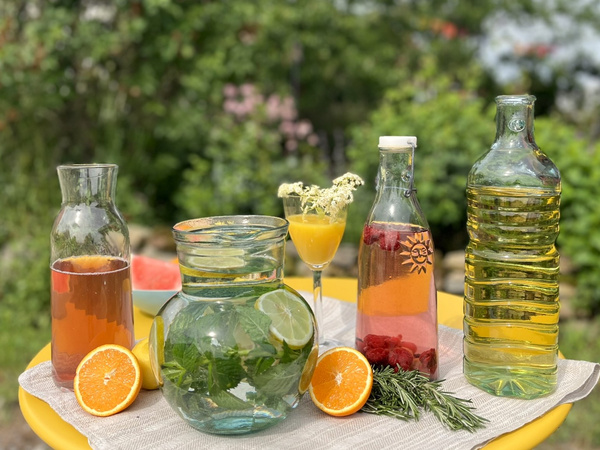 KI generiert: Verschiedene Glasflaschen mit bunten Getränken, frische Früchte und Kräuter auf einem Tisch im Freien.