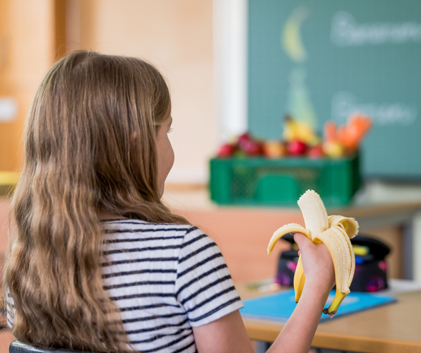KI generiert: Ein Mädchen hält eine Banane in einem Klassenzimmer. Im Hintergrund steht eine Kiste mit Obst.