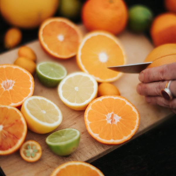 KI generiert: Auf dem Bild sind verschiedene Zitrusfrüchte auf einem Holzschneidebrett zu sehen, darunter Orangen, Zitronen und Limetten, die teilweise aufgeschnitten sind. Eine Hand schneidet mit einem Messer eine Orange.