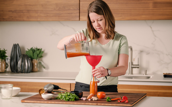 KI generiert: Frau in der Küche füllt Sauce mit einem Trichter in eine Flasche. Zutaten liegen auf dem Tisch.