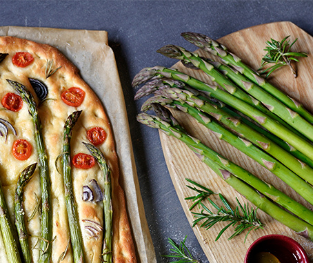 KI generiert: Focaccia mit Spargel und Tomaten auf Holzplatte, daneben frischer grüner Spargel und Rosmarin.