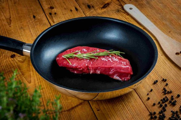 KI generiert: Rotes Steak mit Rosmarin in einer Pfanne auf Holztisch, daneben Pfefferkörner und Holzspatel.