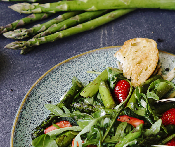 KI generiert: Ein Teller mit Rucolasalat, Erdbeeren, Spargel und Brot auf einem dunklen Hintergrund.