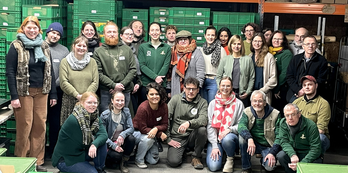 KI generiert: Eine Gruppe von Menschen posiert gemeinsam vor gestapelten grünen Kisten in einem Lagerraum.