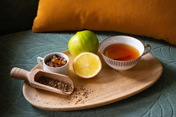 KI generiert: Tee, Zitrone, Kandiszucker und Kräuter auf einem Holztablett vor einem gelben Kissen.