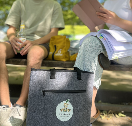 KI generiert: Zwei Personen sitzen auf einer Parkbank. Im Vordergrund ist eine Tasche mit dem Logo "lotta karotta".