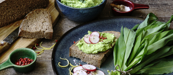 KI generiert: Roggenbrot mit Avocado-Creme und Radieschen, daneben frische Zutaten wie Bärlauch und Zitronenschale.