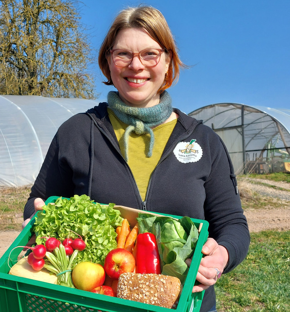 KI generiert: Eine Frau hält eine Kiste mit frischem Gemüse und Obst vor Gewächshäusern. Text auf der Jacke: "lotta karotta".