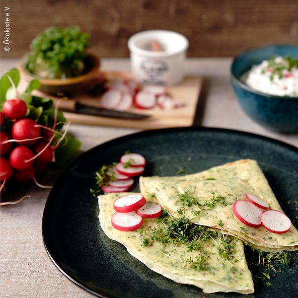 KI generiert: Crepes mit Radieschen und frischen Kräutern, daneben liegen Radieschenbündel. Text: „© Ökokiste e.V.“