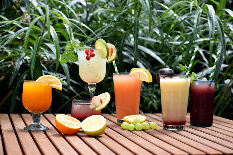 KI generiert: Bunte Fruchtcocktails auf einem Holztisch vor grünen Blättern, dekoriert mit Früchten.