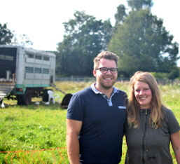 KI generiert: Ein lächelndes Paar steht auf einer grünen Wiese, Hintergrund mit Bäumen und einem Anhänger.