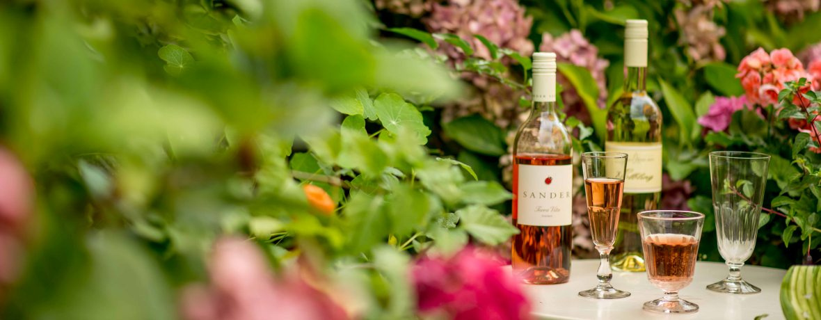 KI generiert: Weinflaschen und Gläser auf einem Tisch in einem blühenden Garten. Text auf der Flasche: "Sandera".