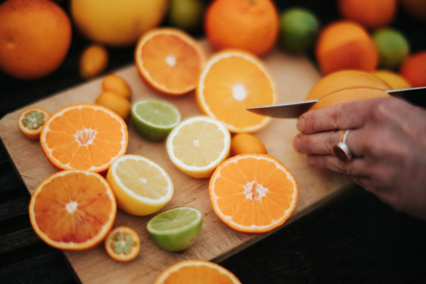 KI generiert: Auf dem Bild sind verschiedene Zitrusfrüchte auf einem Holzschneidebrett zu sehen, darunter Orangen, Zitronen und Limetten, die teilweise aufgeschnitten sind. Eine Hand schneidet mit einem Messer eine Orange.