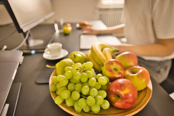 KI generiert: Ein Teller mit frischem Obst auf einem Schreibtisch, im Hintergrund eine Person am Computer.