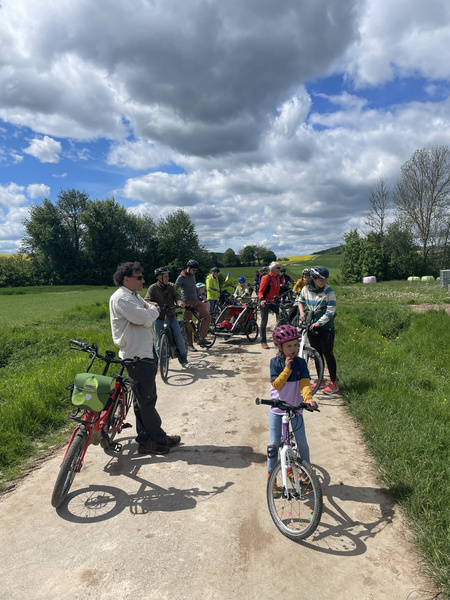 KI generiert: Eine Gruppe von Radfahrern hält auf einem ländlichen Weg an, umgeben von Wiesen und Bäumen unter einem wolkigen Himmel. Einige der Radfahrer unterhalten sich, während ein Kind auf einem Fahrrad im Vordergrund steht.