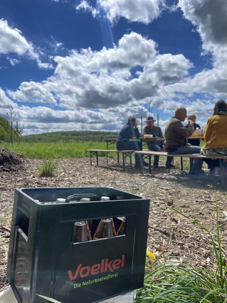 KI generiert: Das Bild zeigt eine Kiste Voelkel-Getränke im Vordergrund und eine kleine Gruppe von Menschen, die draußen an einem Tisch unter einem bewölkten Himmel sitzen. Im Hintergrund erstreckt sich eine grüne Landschaft.