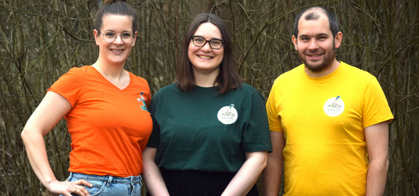 KI generiert: Drei Personen lächeln, tragen bunte T-Shirts mit Logos, im Hintergrund sind Bäume zu sehen.
