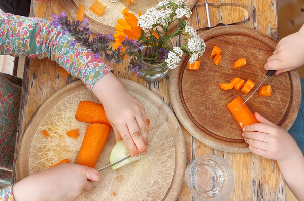 KI generiert: Kinder schneiden Karotten und Zwiebeln auf Holzbrettchen. Ein Blumenstrauß steht dazwischen.