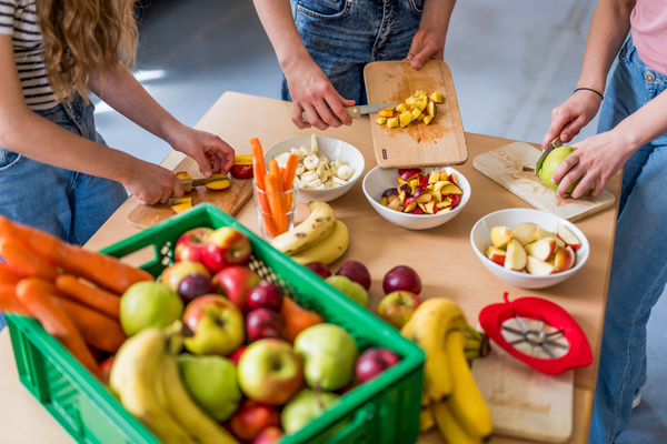 KI generiert: Personen schneiden Obst und Gemüse; bunte Auswahl auf Tisch und in Kiste.